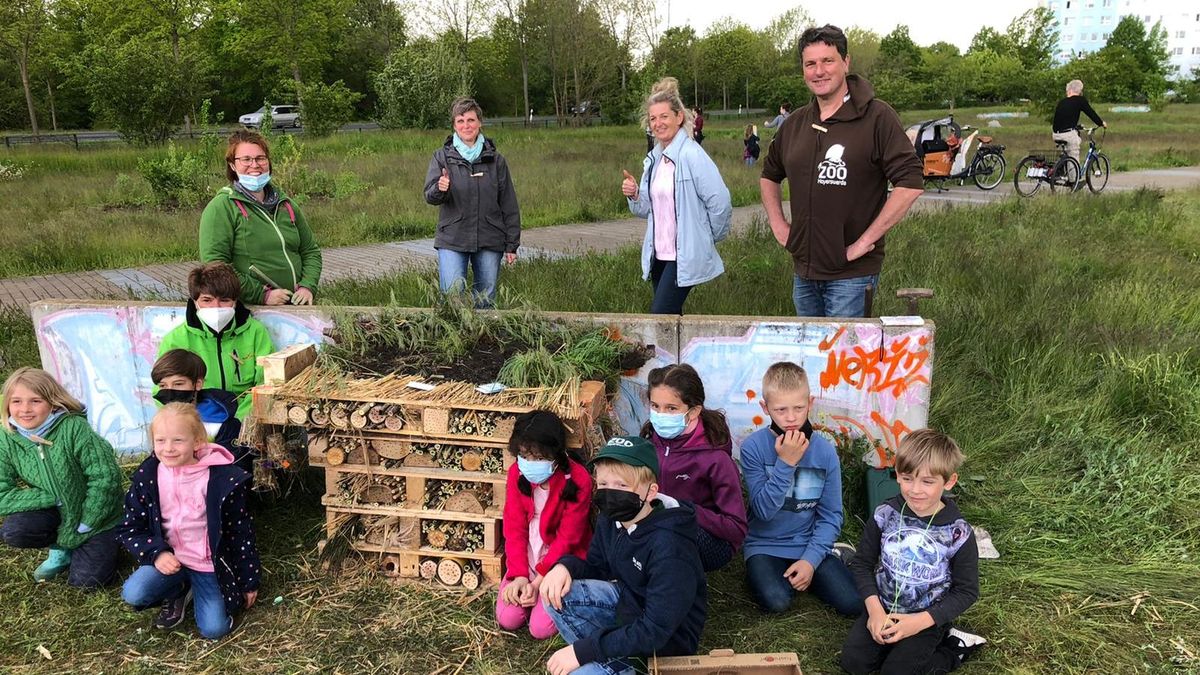 Mehrere Kinder und Erwachsene knien vor einer niedrigen Mauer. Zwischen ihnen sind Paletten mit Holz und Gras gestapelt. Vor ihnen steht eine Kiste mit Heu und Stroh. Hinter der ungefähr kniehohen Mauer stehen vier erwachsene Personen, die in die Kamera lächeln. Um sie herum ist Wiese und ein gepflasterter Weg, sowie ein Hochhaus am rechten Rand des Fotos.  