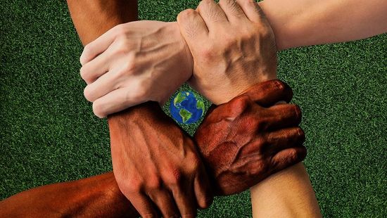 Hände unterschiedlicher Hautfarben greifen einander an den Handgelenken. In ihrer Mitte ist eine Weltkugel.