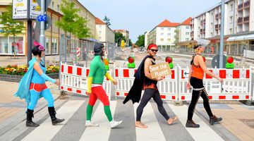 Vier Menschen in Superheld-Kostümen laufen über einen Zebrastreifen. Hinter ihnen ist eine Baustellenabsperrung aufgestellt.