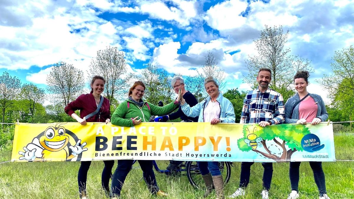 Mehrere Personen halten ein Banner hoch und lachen dabei in die Kamera. Auf dem Banner sind Zeichnungen von Bienen und einem Baum. In weißer, schwarzer und oranger Schrift steht ‘A Place to Beehappy – Bienenfreundliche Stadt Hoyerswerda’. Im Hintergrund ist noch eine Person auf einem Fahrrad zu sehen. Die Personen stehen auf einem Feld mit Bäumen dahinter. 