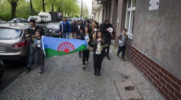  Eine Gruppe Erwachsener und Jugendlicher laufen auf die Kamera zu. Es wirkt als wären sie auf einem Straßenumzug. Ganz vorne halten zwei Jugendliche eine die Flagge der Roma, diese ist grün und blau. In der Mitte der Fahne ist ein halbes, rotes Rad mit Ritzen zu sehen. Links neben den Menschen stehen Autos.  