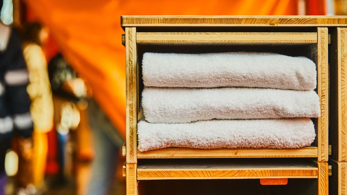 Im rechten Bereich des Bildes sind drei weiße Handtücher in einem Regal gestapelt. Im Hintergrund ist ein oranges Tuch zu sehen. 