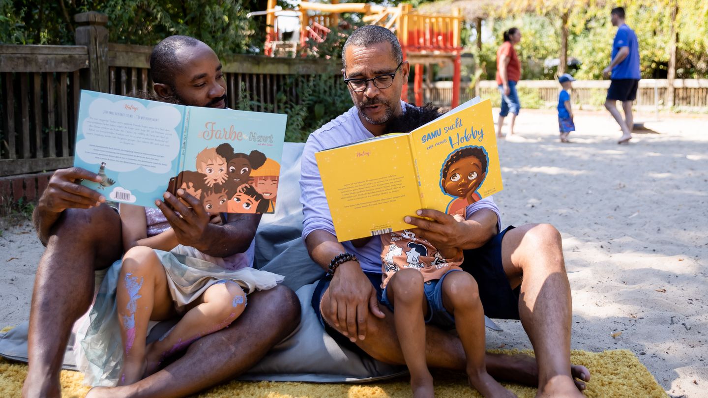Zeigt bei Männer von BLACK DADS READERS mit jeweils einen aufgeschlagenen Buch in der Hand. Auf ihrem Schoss sitzen zwei Kinder, denen sie das Buch vorlesen.  In der oberen Hälfte des Bildes stehen 2 Personen  und ein Kind