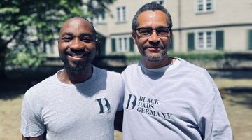 Zwei Schwarze Menschen schauen lächelnd in die Kamera. Sie haben beide ein Hellgraues Shirt an mit dem Logo des Vereins: BDG – BLACK DADS GERMANY.