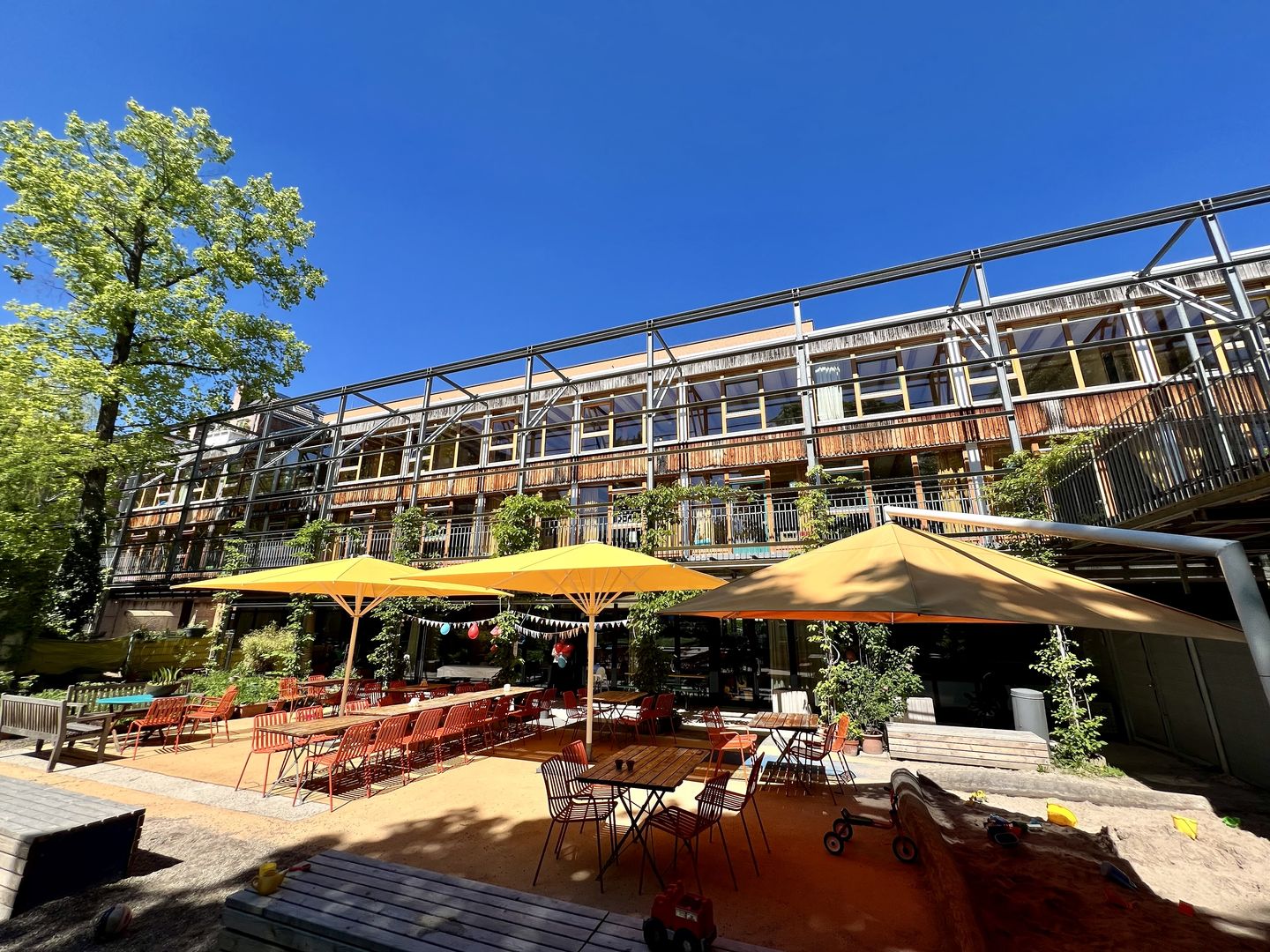 Es ist die einladende Gartenterrasse des EKiZ unter gelben Sonnenschirmen und blauem Himmel zu sehen. 