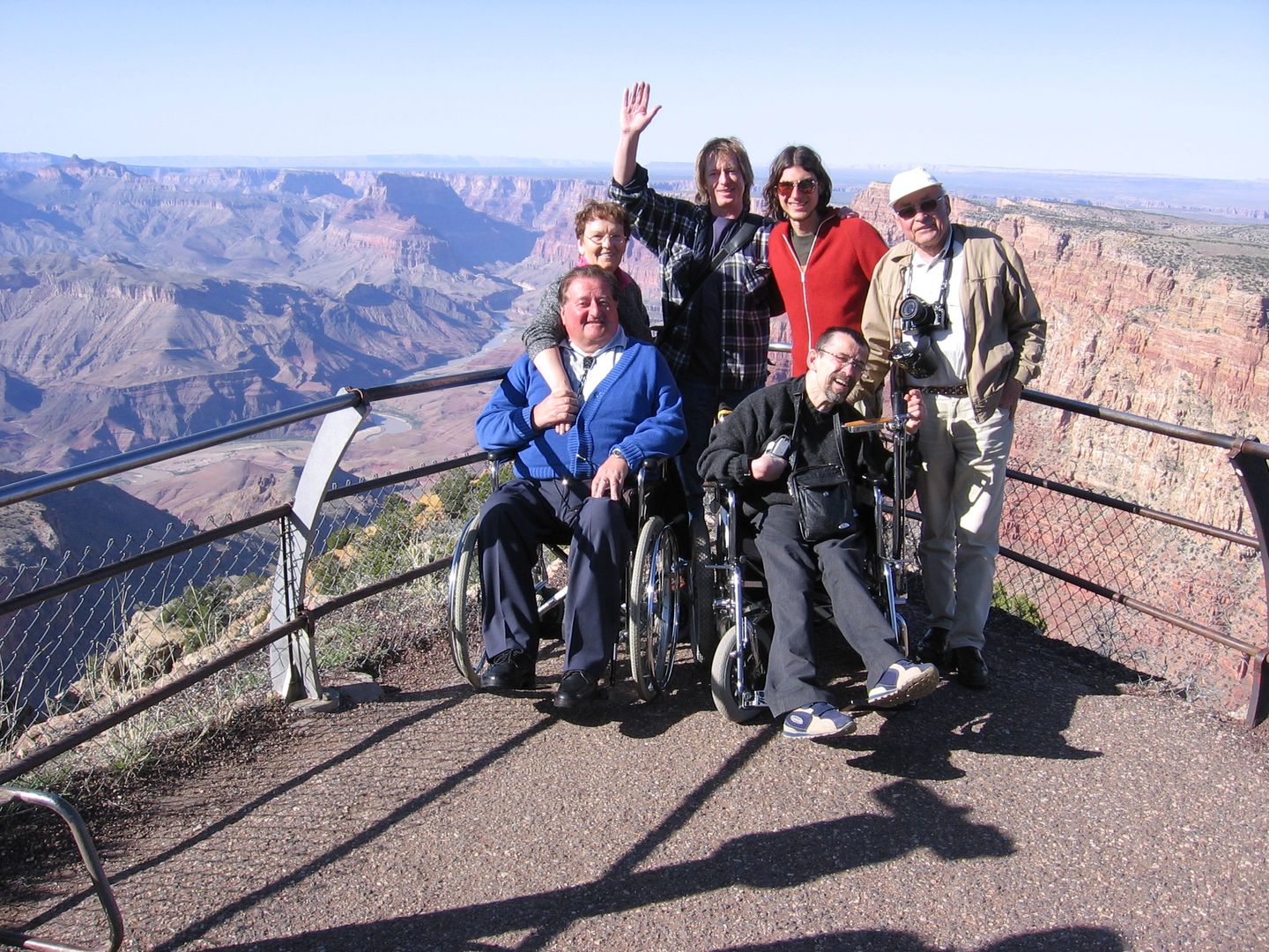 Eine Reisegruppe von 6 Personen stehen an einem Aussichtspunkt auf den Gran Canyon. zwei Rollstuhlfahrer*innen sind dabei. Alle winken gut gelaunt in die Kamera. Die Sonne scheint. 