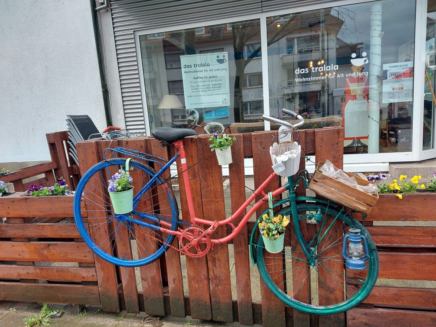 Es ist eine Aussenansicht vom Cafe tralala zu sehen. An einem Zaun aus Paletten hängt ein mit Blumen geschmücktes Fahrrad