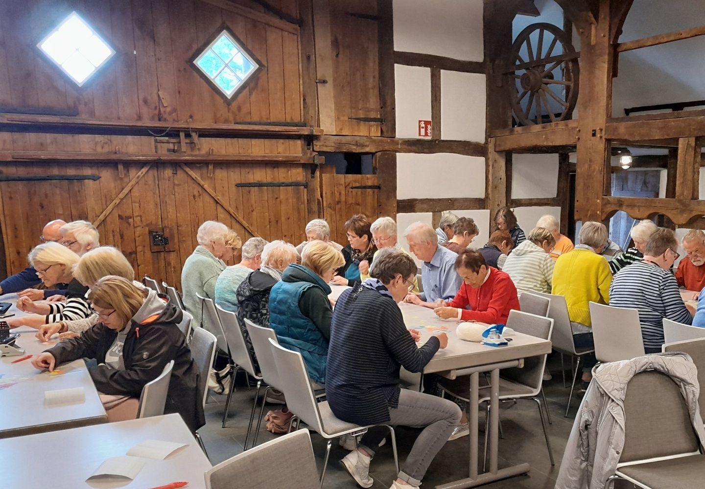 Auf dem Foto sitzen Personen an Tischen, überwiegend mit dem Rücken zum Fotografen. Ihre Köpfe sind nach unten mit Blick auf die Tischplatte geneigt.  Sie malen oder kleben etwas auf Papier.