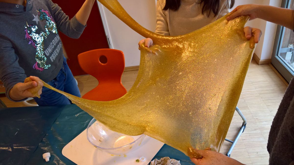 In der Mitte des Fotos ist eine gold-glitzernde Slime-Masse zu sehen. Sie wird an den Ecken von mehreren Händen auseinandergezogen und erinnert an ein aufgespanntes Tuch.  Im Hintergrund stehen ein oranger Stuhl sowie eine Glasschüssel.  