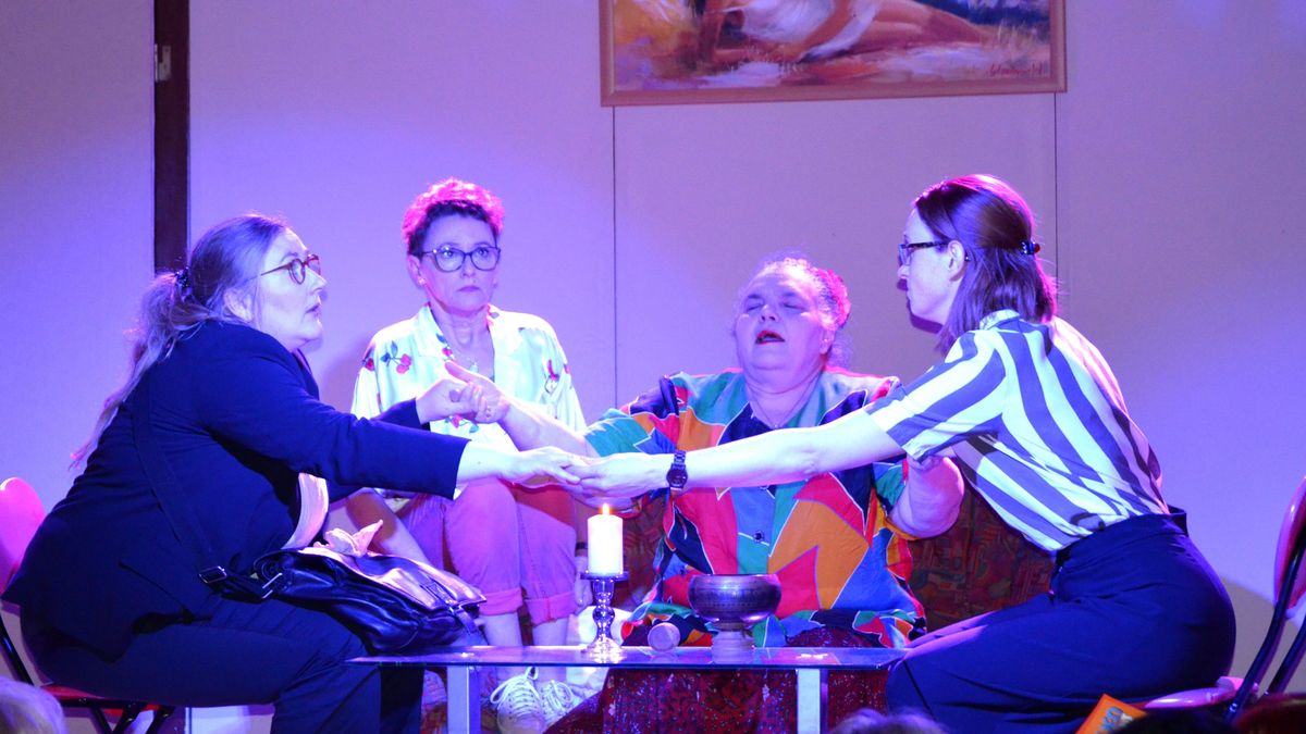 Auf einer Bühne sitzen vier Frauen in einem Wohnzimmer. Drei Frauen reichen sich die Hand. Die vierte Frau sitzt daneben. Über ihnen hängt ein Gemälde an der Wand.