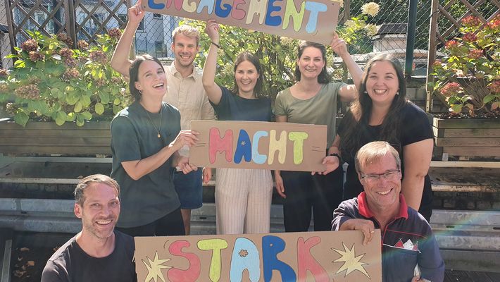 Das Engagementteam der Verbraucherzentrale, 7 Personen, lächeln und hälten Schilder hoch mit der Aufschrift "Engagement macht stark"