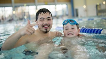 Zwei Menschen mit Down-Syndrom - ein Mann und ein Kind - befinden sich in einem Schwimmbecken in einem Hallenbad und lächeln in die Kamera.
