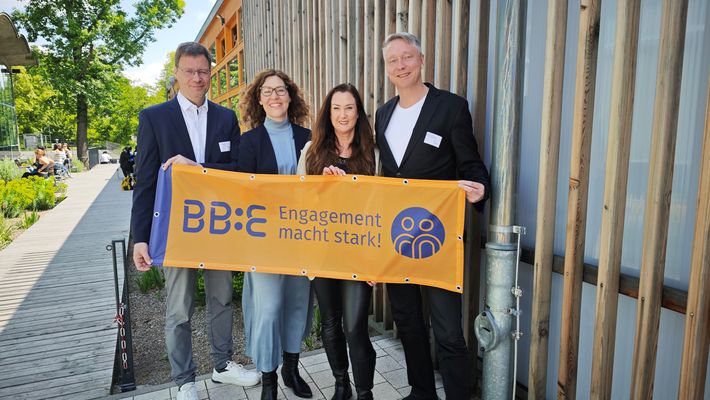 Vier Personen – zwei Männer und zwei Frauen – stehen lächelnd nebeneinander. Gemeinsam halten sie ein orangefarbenes Banner mit dem BB:E-Logo und dem Schriftzug „Engagement macht stark!"