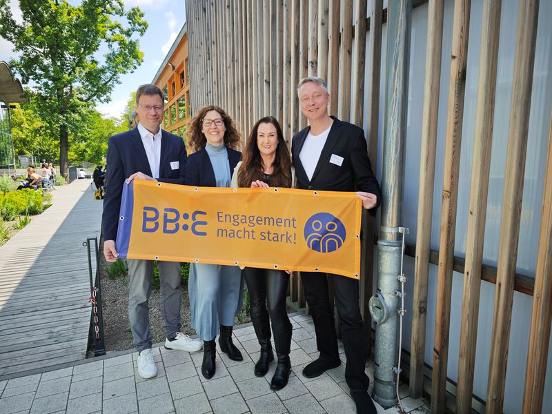 Vier Personen – zwei Männer und zwei Frauen – stehen lächelnd nebeneinander. Gemeinsam halten sie ein orangefarbenes Banner mit dem BB:E-Logo und dem Schriftzug „Engagement macht stark!"
