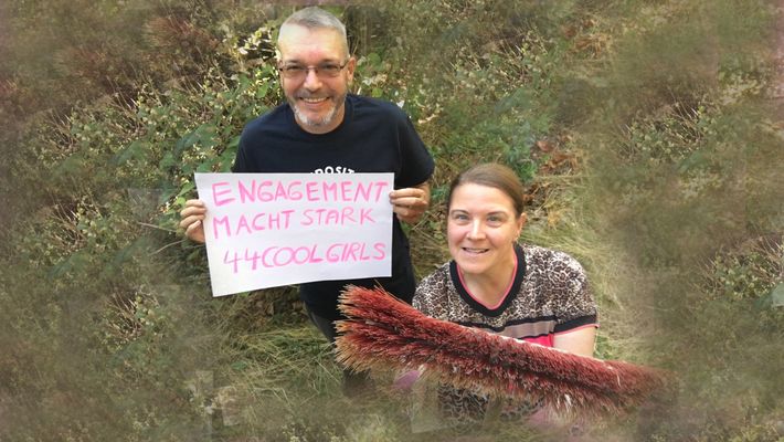 Zwei Menschen stehen auf einer Wiese. Eine Person hält ein Schild, die andere Person einen Besen in der Hand.