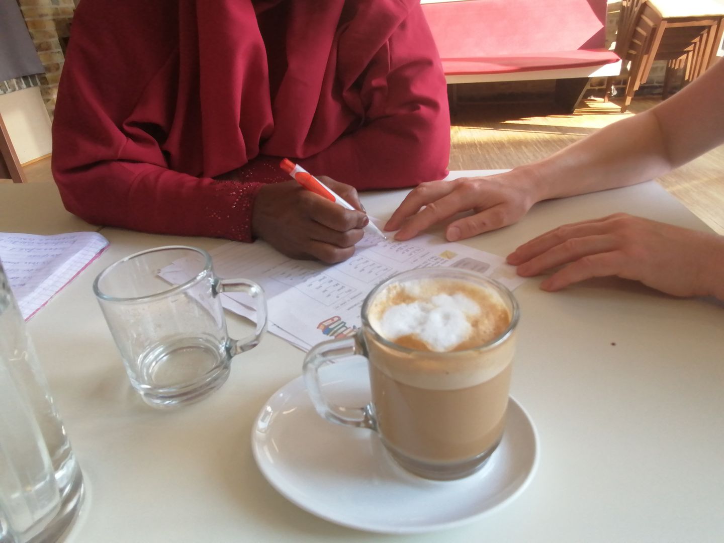 Hände von zwei Frauen, eine schreibt, daneben eine Tasse Cappucino