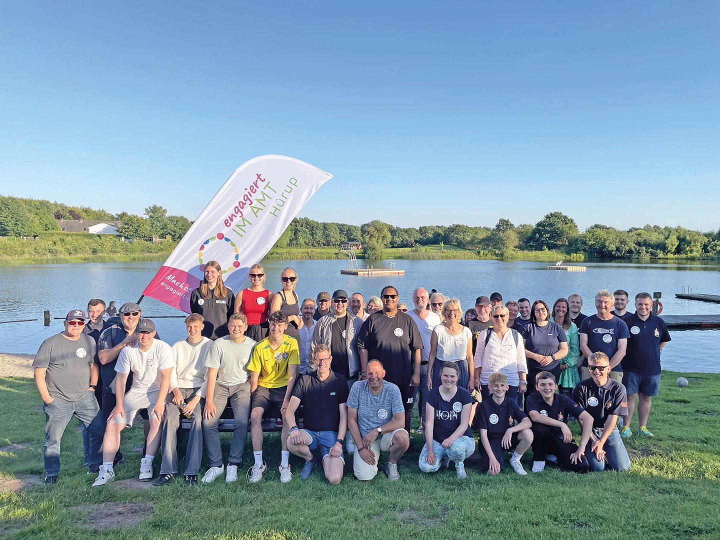Auf dem Bild ist eine Gruppe von ca. 40 Personen zu sehen, die alle etwas gemeinsam haben: Sie sind ehrenamtlich engagiert. Sie stehen bei sehr schönem Wetter fröhlich beisammen und im Hintergrund ist eine Badesee zu sehen. 