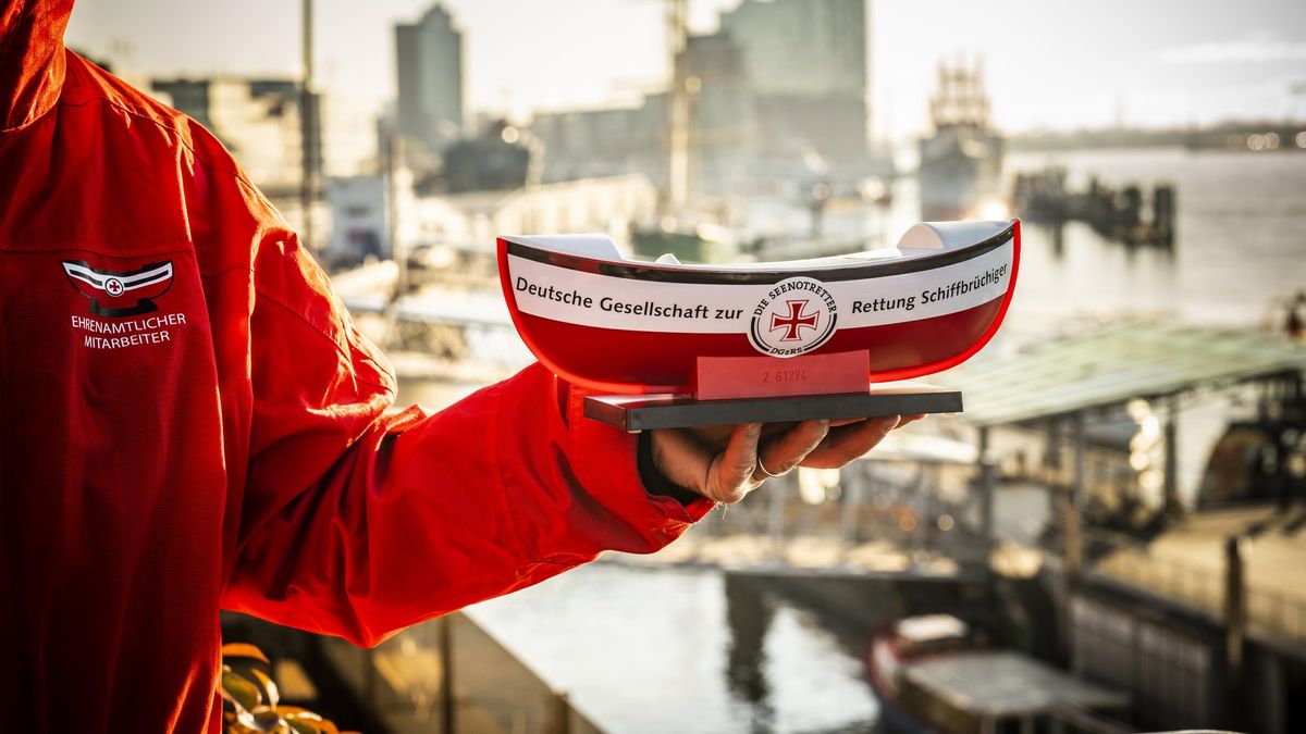 Eine Person hält ein rotes Schiffchen in der Hand, auf dem in schwarzer Schrift steht „Deutsche Gesellschaft zur Rettung Schiffbrüchiger“. In der Mitte befindet sich das Logo der DGzRS. Im Hintergrund kann man Hamburg erkennen. 
