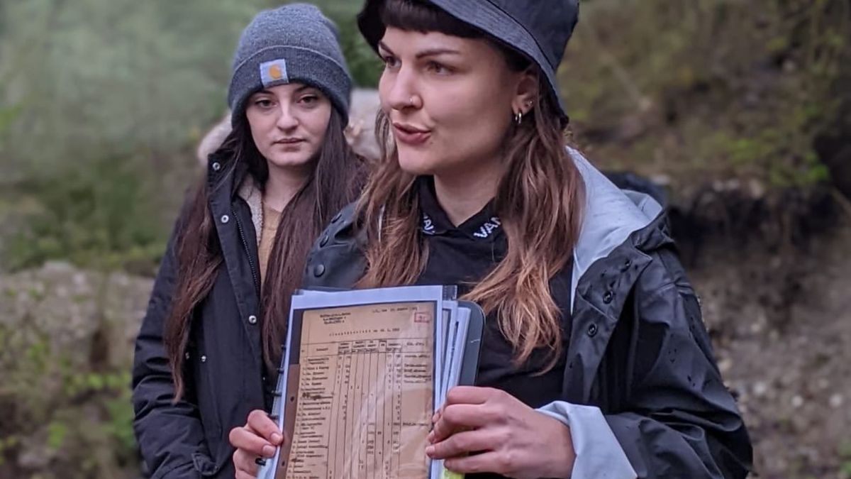 Zu erkennen sind Susanne Siegert und eine Frau, die links neben ihr steht. Susanne Siegert hält in der Hand Dokumente. Der Hintergrund ist verschwommen und man kann einen Wald erkennen.