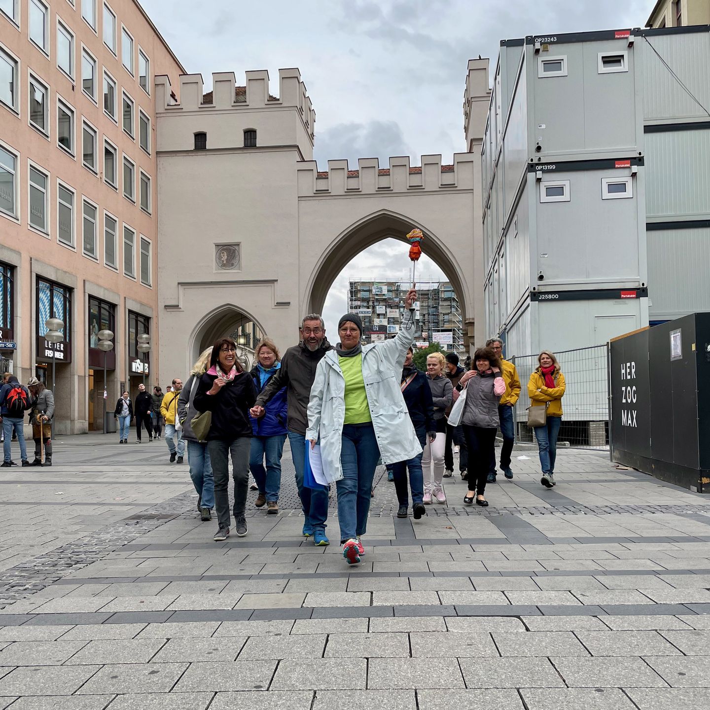 Das Foto zeigt ein eGruppe von Menschen, die durch die Münchner Innenstadt geht. Voran geht ein eFrau, di einene Schirm nach oben hält, damit die Teilnehmenden des Stadtspaziergangs sie sehen können. 