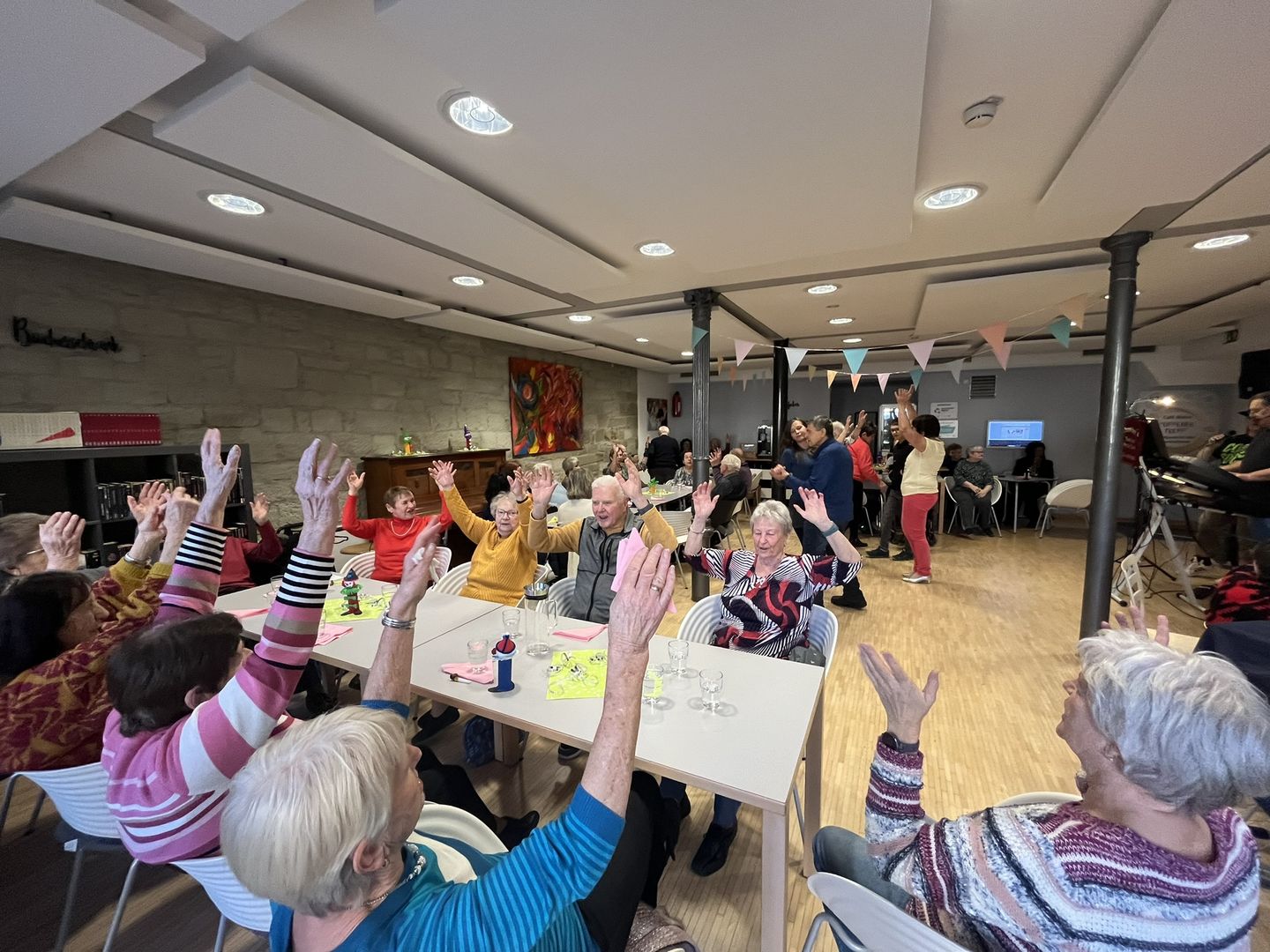 Viele Menschen sitzen an einem Tisch und schunkeln, einige Menschen tanzen im Raum.