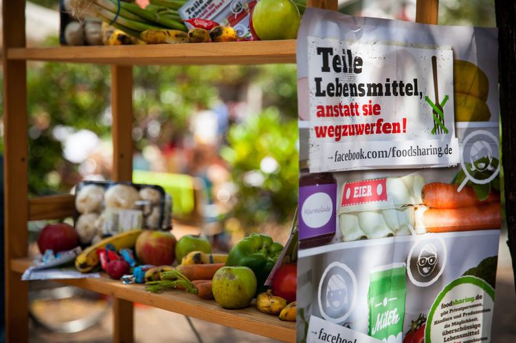 Auf einem Holzregal liegen Obst und Gemüse. An der Seite klebt ein Plakat: Teile Lebensmittel, anstatt sie wegzuwerfen.