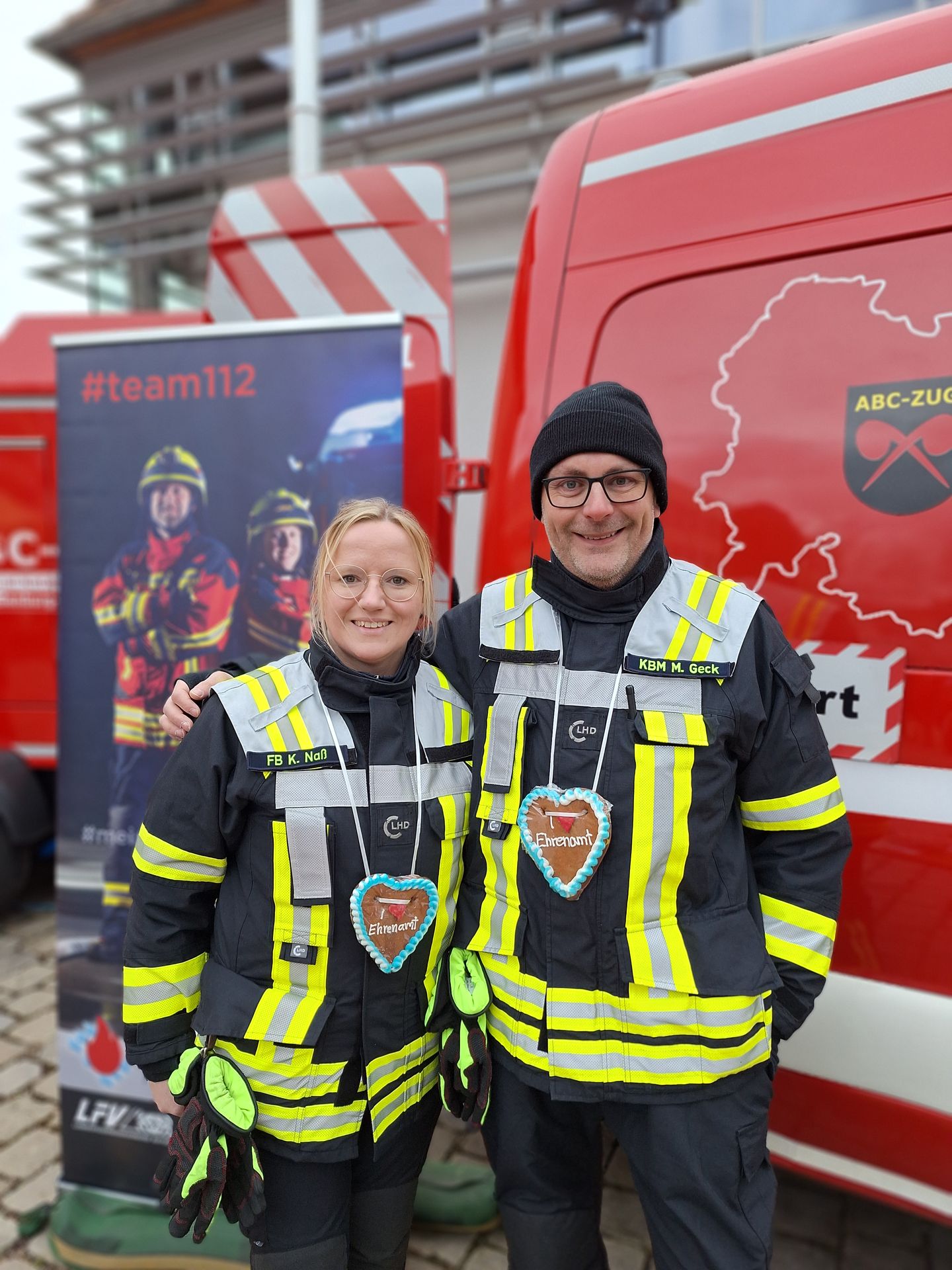 Zwei Feuerwehrleute, ein Mann und eine Frau, stehen in Schutzanzügen vor einem Feuerwehrauto