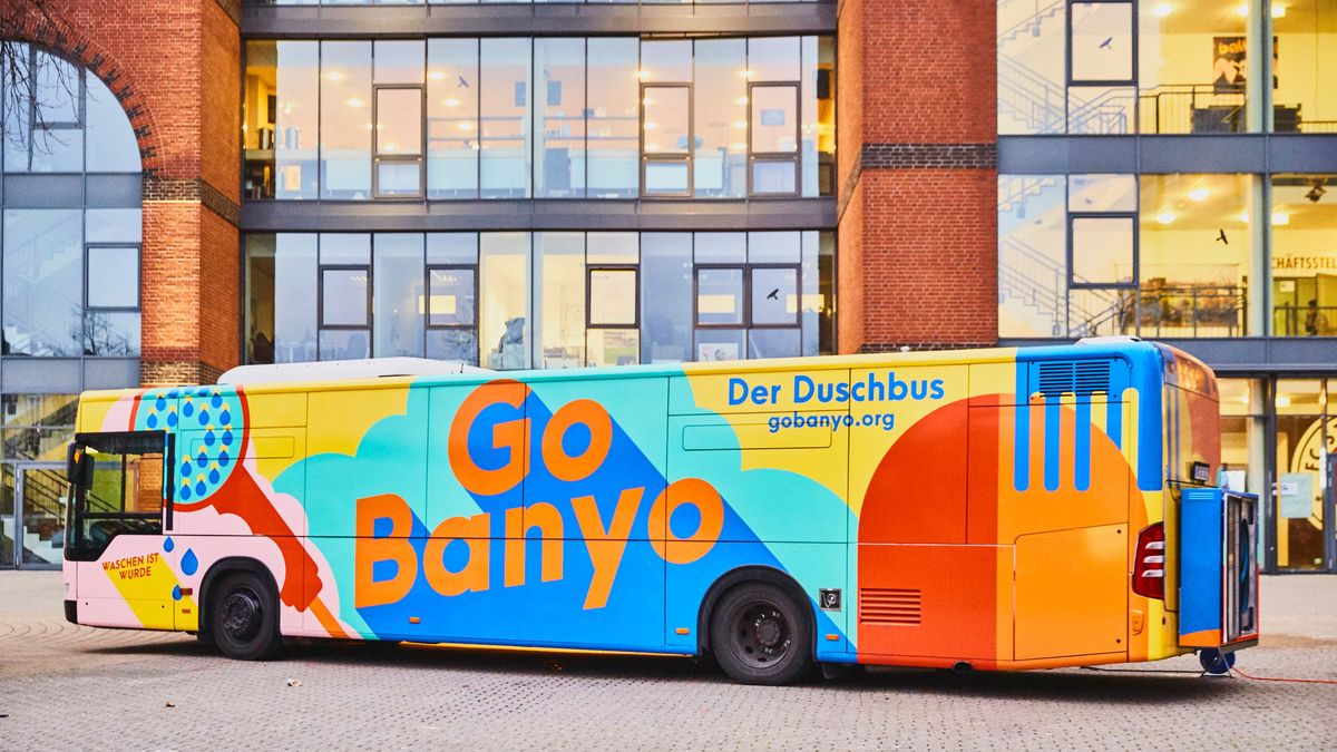Ein bunter Linien-Bus auf dem groß ‚Go Banyo‘ und ‚Der Duschbus – gobanyo.org‘ steht. Der Bus steht vor einem Gebäude mit beleuchteten Glasfenstern.  