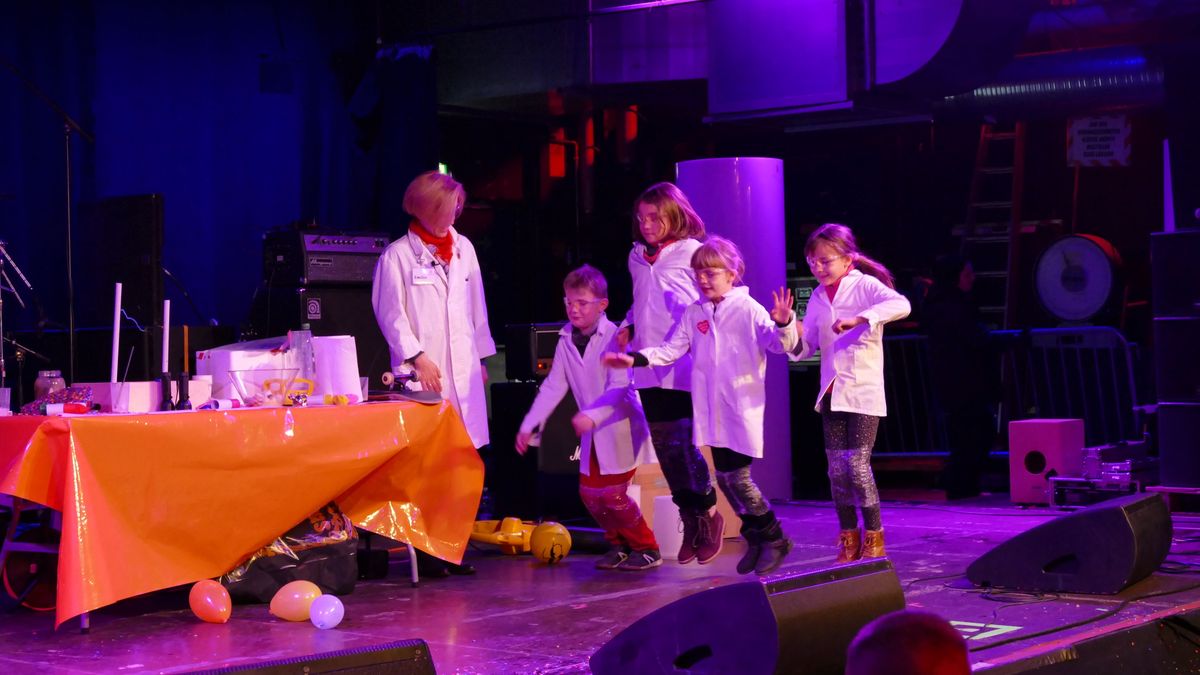Mehrere Kinder in weißen Labor-Kitteln und mit Schutzbrillen laufen auf einer Bühne auf einen Tisch mit einer orangen Tischdecke zu. Neben dem Tisch steht eine Frau, ebenfalls in einem weißen Laborkittel. Auf dem Tisch sind verschieden Gegenstände für einen Versuch aufgebaut.