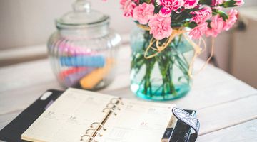 Aufgeschlagener Terminkalender und eine Glasvase mit rosa Nelken auf einem weißen Holztisch.