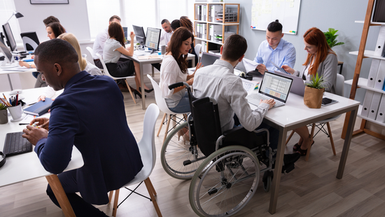 Ein Raum mit mehreren Tischen, an denen teilweise PCs und Laptops stehen. An den Tischen sitzen junge Menschen unterschiedlichen Geschlechts und unterschiedlicher Hautfarbe sowie eine Person im Rollstuhl. Teilweise schauen sie auf ihre Arbeitsgeräte, teilweise diskutieren sie miteinander. 