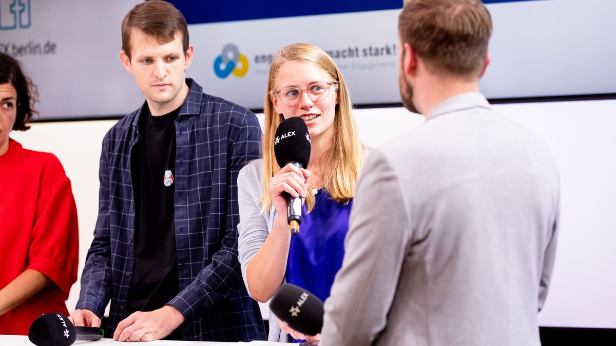 Henrike Schlottmann von der Project Together gGmbH spricht in das Mikro, links von ihr stehen Markus Fleige und Elena Kountidou. Rechts mit dem Rücken zur Kamera steht der Moderator. 