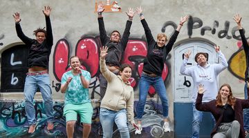 Junge Menschen springen vor einer Graffitiwand in die Luft.