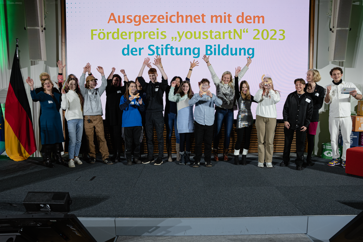 [ALT: Ein Foto mit einer Bühne voller junger Menschen bei einer Preisverleihung. Über ihnen steht in bunten Buchstaben der Text Ausgezeichnet mit dem Förderpreis youstartN 2023 der Stiftung Bildung]  