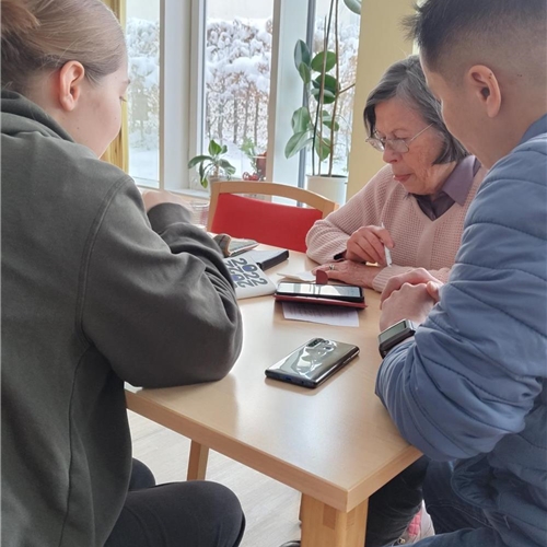 Zwei junge Menschen sitzen mit einer ältereren Frau zusammen und führen ein Beratungsgespräch rund um ihr Smartphone durch.