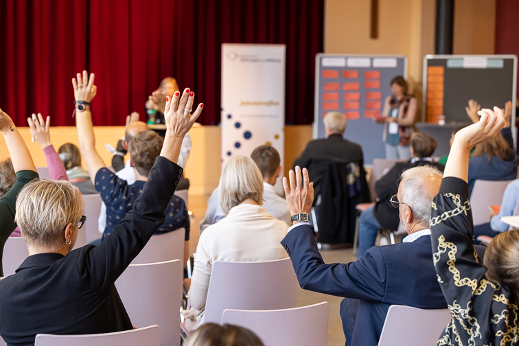 Teilnehmende des Jahrestreffens 2022 sitzen im Plenumssaal. Im Hintergrund steht die Moderatorin. Teilnehmende heben die Hand zur laufenden Abstimmung.