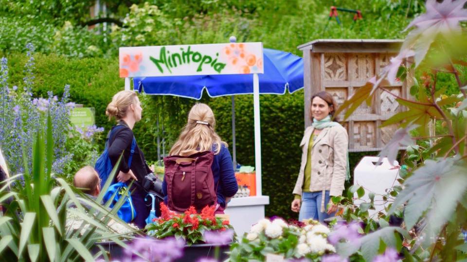 ein Garten mit Grünpflanzen und Blumen, drei Personen sind im Garten, sie gucken sich an, im Hintergrund ist eine Bude mit MArkise darauf der Schriftzug "minitopia"