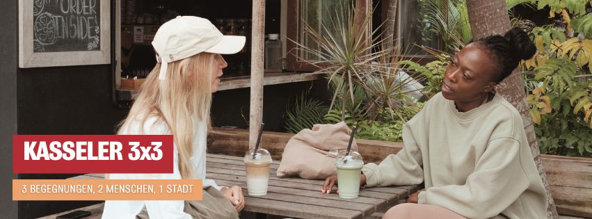 Zwei Frauen sitzen sich draußen an einem Holztisch gegenüber und unterhalten sich bei einem Getränk. Eine trägt ein helles Basecap und eine weiße Bluse, die andere ein beigefarbenes Sweatshirt. Sie wirken konzentriert und zugewandt. Im Hintergrund sind Pflanzen und ein Caféfenster zu sehen. Das Bild steht für eine offene, persönliche Begegnung im Projekt Kasseler 3x3.