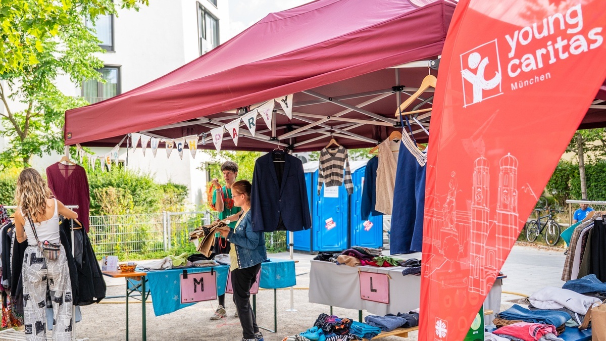 In einem Pavillon ist auf Tischen Kleidung in verschiedenen Größen sortiert. 3 junge Menschen stöbern durch das Angebot.