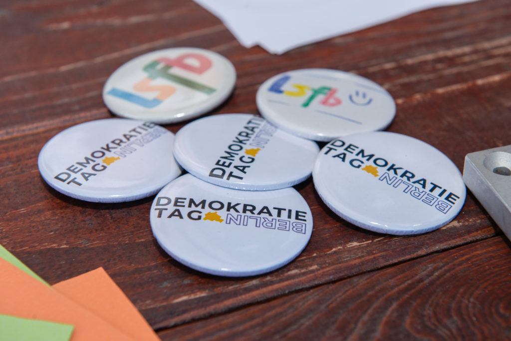 sechs Ansteckbuttons, davon vier mit dem Aufdruck Berliner Demokratietag und zwei mit dem Logo des lsfb