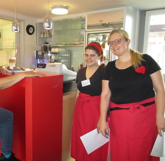 Zwei damen in schwarzen T-Shirts und roten Schürzen stehen in einem Café. Dort sind sie ehrenamtlich tätig. 