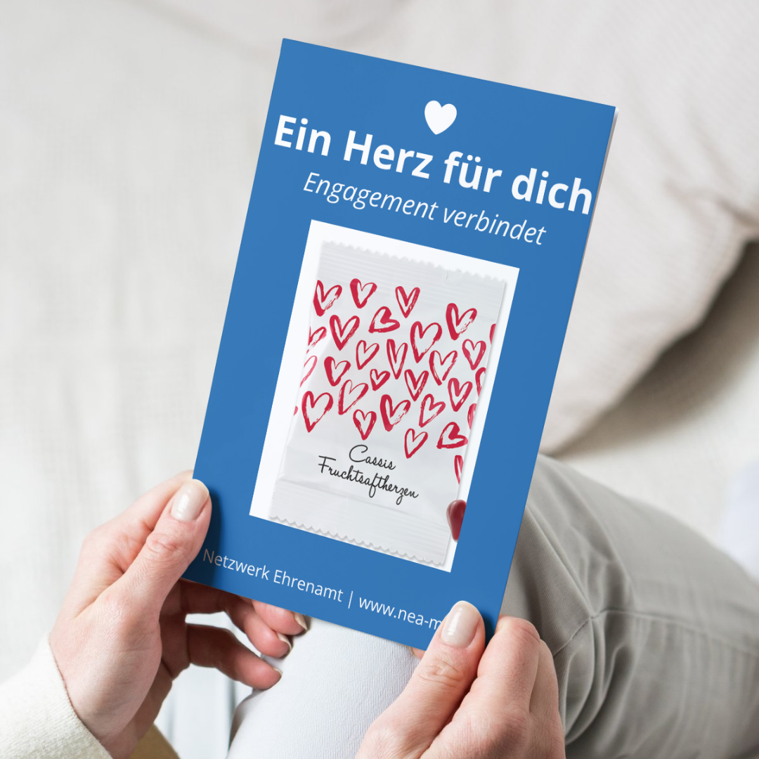 Eine Person hält einen Flyer mit einem blauen Hintergrund in den Händen. Auf dem Flyer steht oben: „Ein Herz für dich – Engagement verbindet“. In der Mitte ist ein kleines Tütchen mit vielen roten Herzmotiven zu sehen, beschriftet mit „Cassis Fruchtsaftherzen“. Unten auf dem Flyer steht: „Netzwerk Ehrenamt | www.nea-mb.de