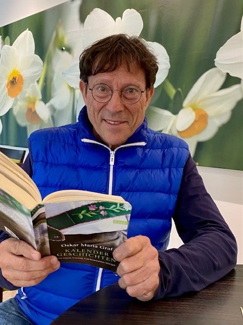 R.V. sitzt uns zugewandt, ein Buch von Oskar Maria Graf in der Hand. Er trägt eine runde Brille und blickt freundlich in die Kamera.