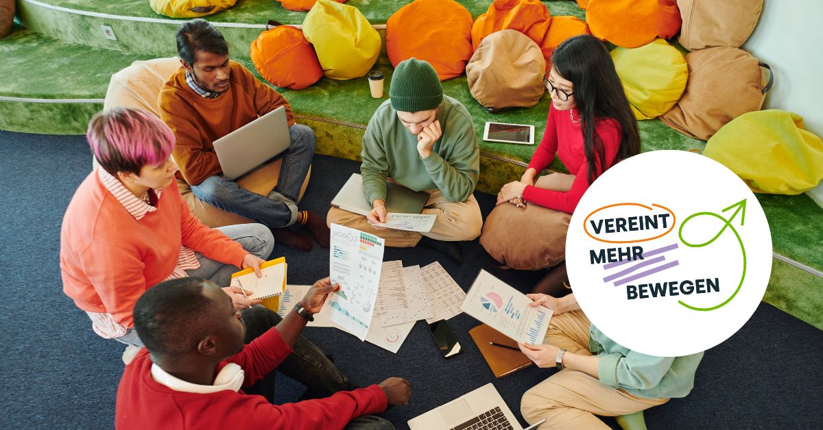 Mehrere Personen sitzen auf dem Boden im Kreis und besprechen sich. Sie halten Unterlagen in den Händen.