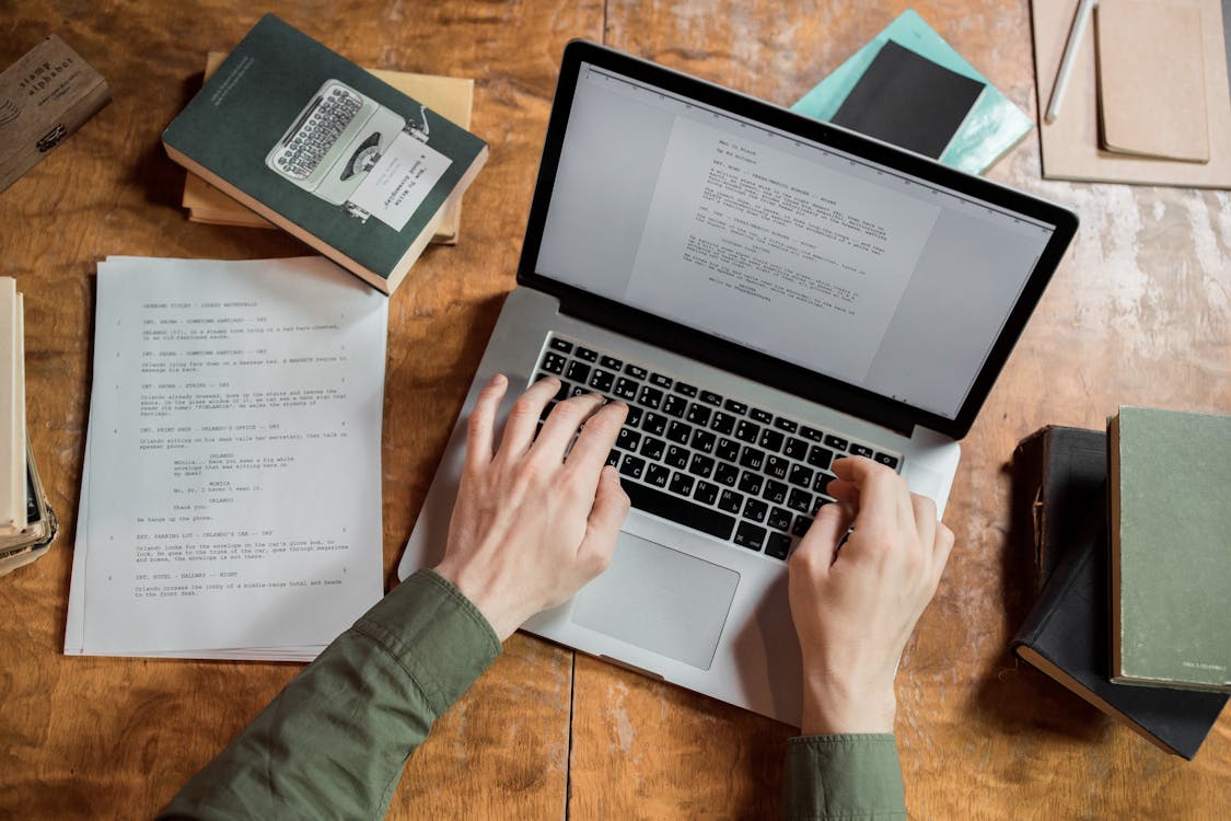 Von oben ist ein Laptop zu sehen an dem zwei Arme arbeiten, also etwas tippen. Drumherum liegen Blätter und ein Notizbuch sowie ein Stift. 